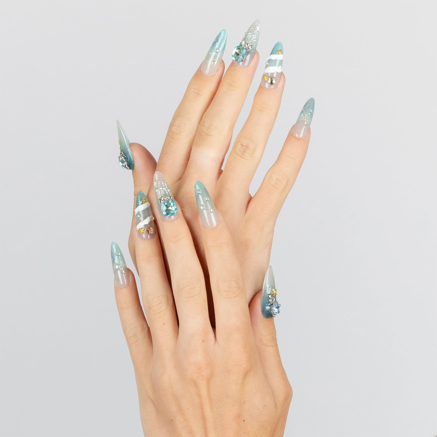 A pair of hands with long, sculpted almond-shaped nails decorated with light blue glitter, white stripes, and small gold and blue rhinestone accents, shown against a plain light gray background.