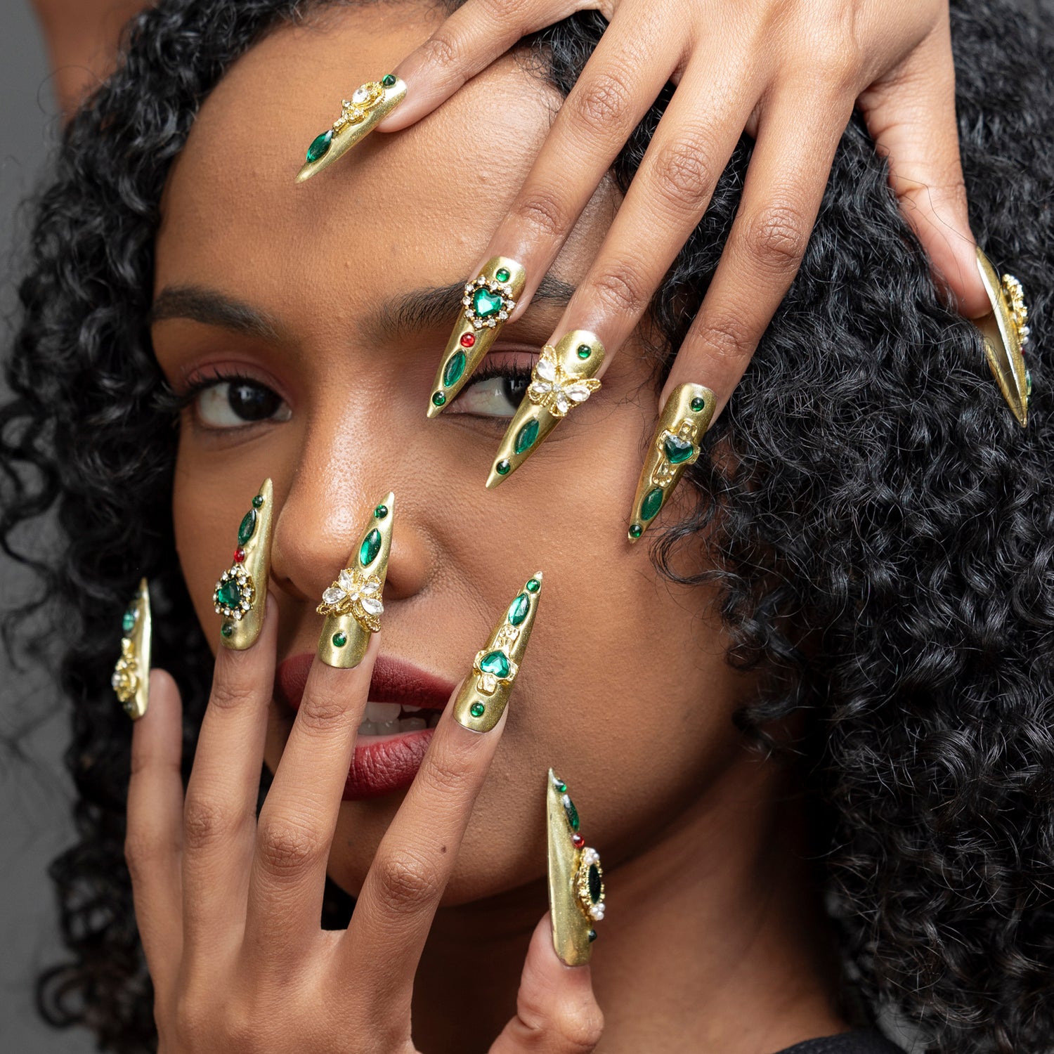 A close-up of a model with long, dramatic stiletto-shaped nails decorated with gold metal, green gemstones, and white floral details, posed against her face.