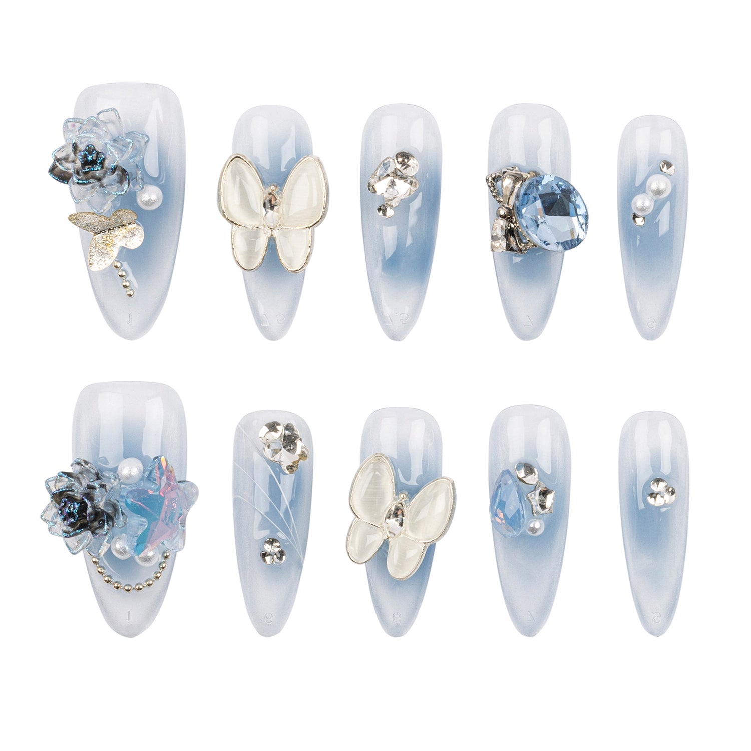Ten false press-on nails arranged in two rows of five on a plain white background. The nails have a pale icy blue to white ombré finish and are decorated with various embellishments including white butterflies, blue crystals, pearls, and metallic charms.