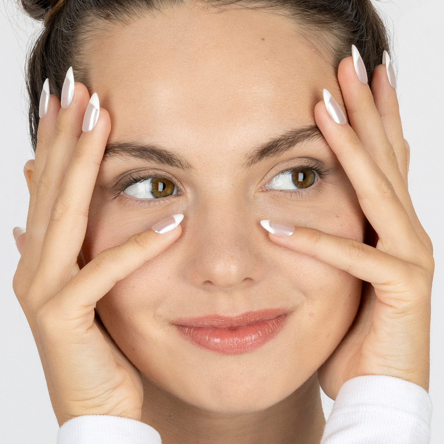 Close-up of a woman with long white French-tip press-on nails, gently touching her cheeks and eyes, wearing a white long-sleeve top against a light gray background.