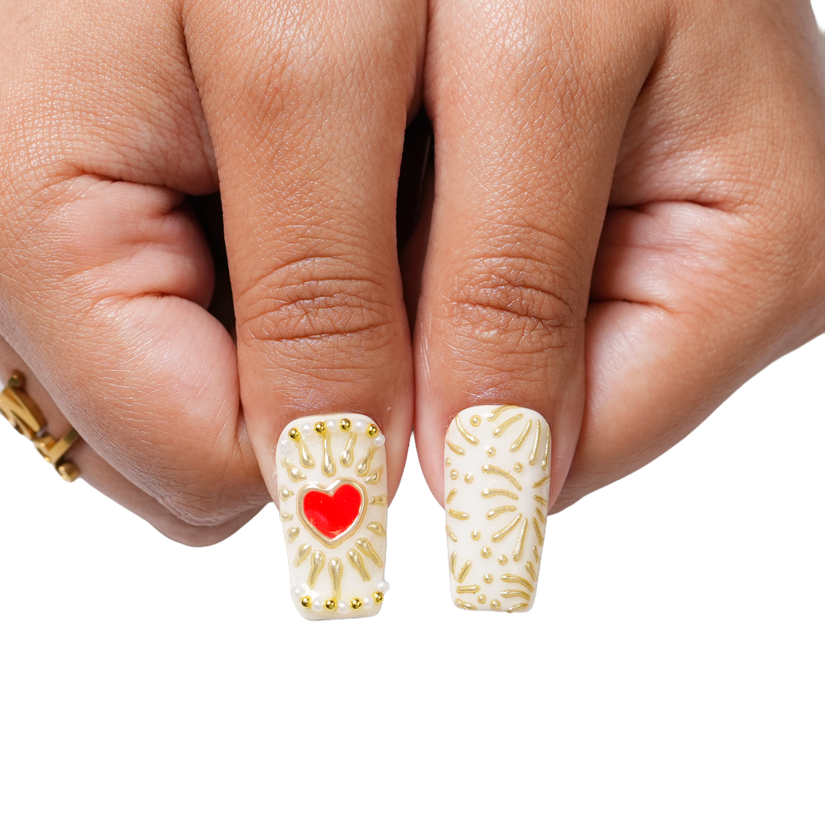 Close-up of Gilded Relic long square press-on nails with ornate gold detailing, pearl-like beads, red and clear gems, and cameo-inspired accents.