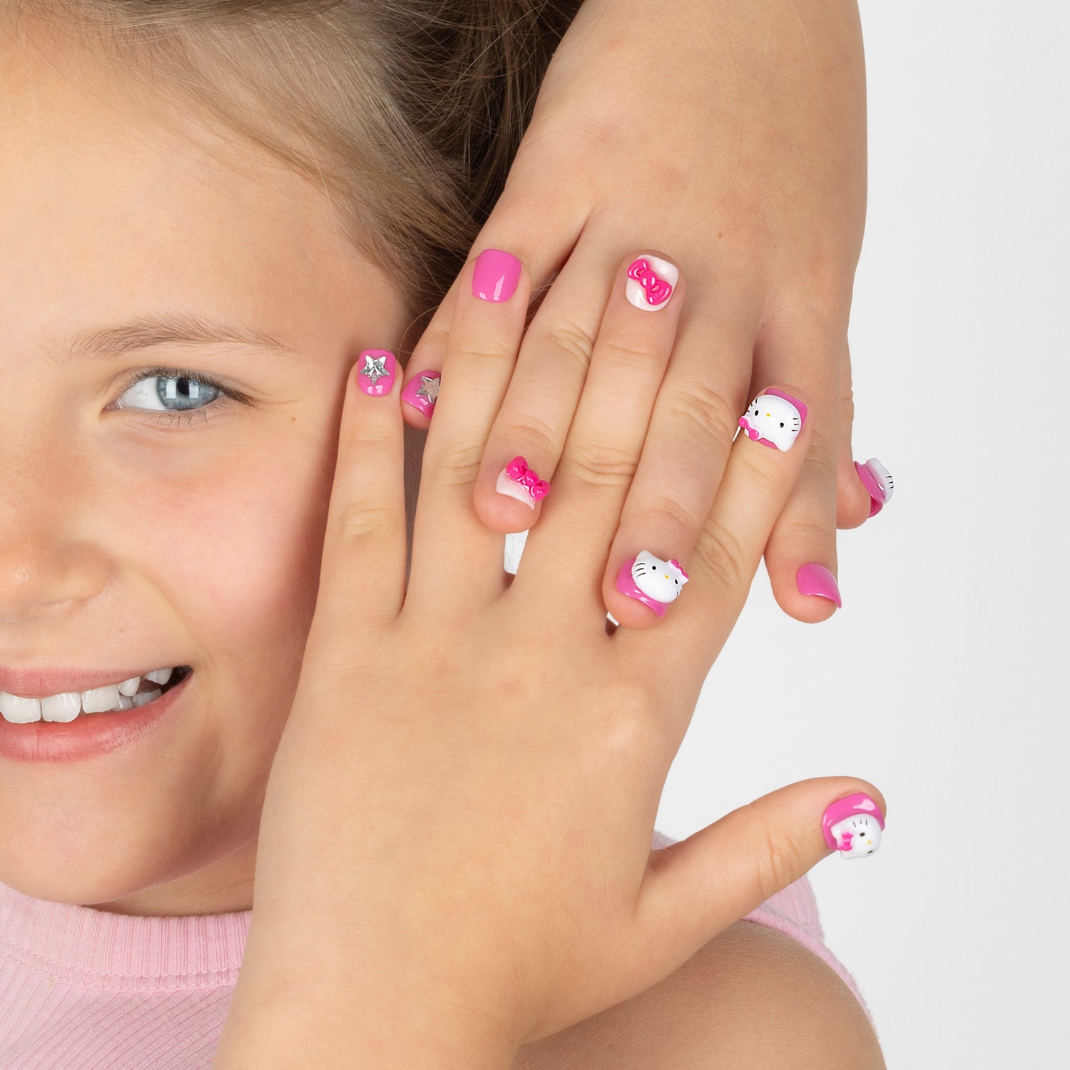 A close-up of a child's hands with pink and white Hello Kitty nail art, with part of the child's face visible on the left against a light gray background.