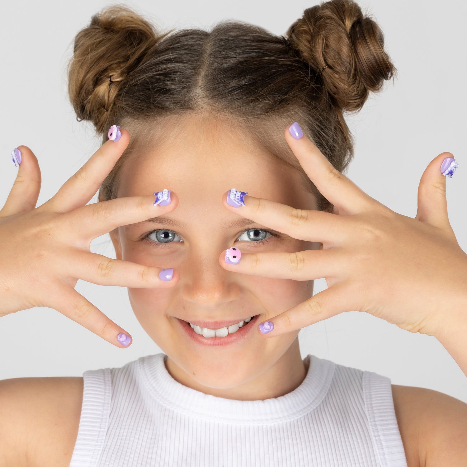 A smiling young girl with her hair in two buns poses with her hands over her eyes, showcasing cute lavender and white Sanrio Kuromi cartoon nail art. She is wearing a white ribbed tank top against a solid light gray background.
