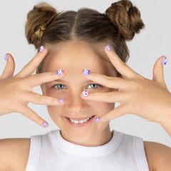 A smiling young girl with her hair in two buns poses with her hands over her eyes, showcasing cute lavender and white Sanrio Kuromi cartoon nail art. She is wearing a white ribbed tank top against a solid light gray background.