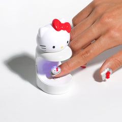 A white Hello Kitty nail lamp with a red bow sits on a white surface while a hand places a decorated nail under the UV light. The nail lamp features the Hello Kitty face design with a red bow, and the hand shows a silver glittery nail with a small red bow and Hello Kitty design.