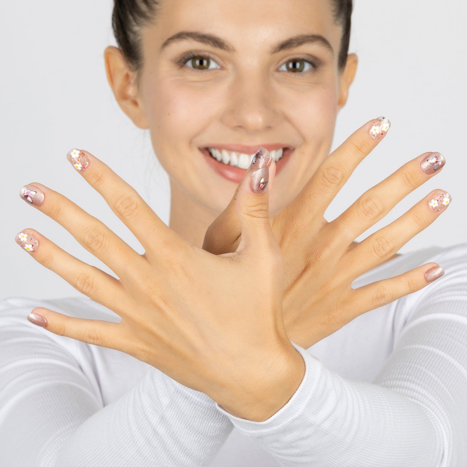 A smiling woman with shimmery pink press-on nails crossed in front of her face, wearing a white long-sleeve top against a light gray background.