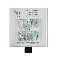 A boxed set of reusable luxury press-on nails. The white square package shows a clear window with ten long light blue and silver decorative nails, plus text reading 'HIGH QUALITY HAND MADE HASSLE FREE' and 'REUSABLE LUXURY NAILS.