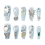 Ten decorative false fingernails arranged in two rows on a plain white background. The nails are light icy blue with white floral and metallic embellishments, including roses, butterflies, bows, rhinestones, and a green gem accent.