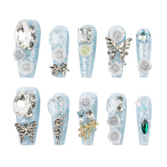 Ten decorative false fingernails arranged in two rows on a plain white background. The nails are light icy blue with white floral and metallic embellishments, including roses, butterflies, bows, rhinestones, and a green gem accent.