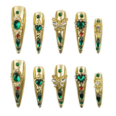 Ten gold stiletto-shaped press-on nails arranged in two rows on a white background, featuring various green, red, and clear rhinestone embellishments.