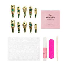 A nail art kit on a white background featuring ten decorated false nails with gold glitter and green gems, a pink alcohol pad packet, a white nail file, a pink nail polish tube, a wooden cuticle stick, and a white stamping plate.