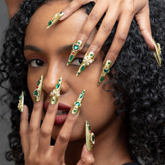 A close-up of a model with long, dramatic stiletto-shaped nails decorated with gold metal, green gemstones, and white floral details, posed against her face.