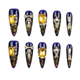 Ten decorative press-on nails arranged in two rows on a plain white background. The nails are elongated stiletto-shaped with a glossy deep blue base, gold glitter detailing, and various yellow gemstone and crystal embellishments.