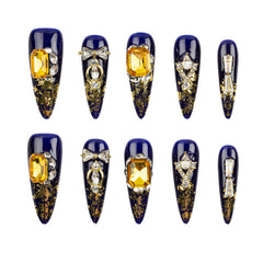 Ten decorative press-on nails arranged in two rows on a plain white background. The nails are elongated stiletto-shaped with a glossy deep blue base, gold glitter detailing, and various yellow gemstone and crystal embellishments.