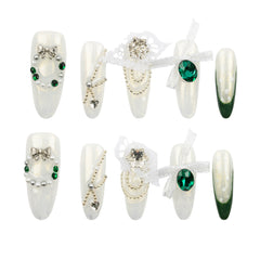 Eight decorative false press-on nails arranged in two rows on a plain white background. The nails are primarily glossy white with embellishments including green gemstones, silver rhinestones, bows, lace, and dark green gradient tips on some pieces.