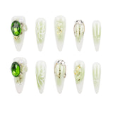 Ten artificial press-on nails arranged in two rows of five on a plain white background. The nails are elongated almond-shaped with a white base and decorative designs including a large green oval stone, white floral swirls, gold glitter, pearl accents, and silver chain details.
