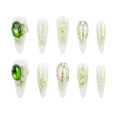 Ten artificial press-on nails arranged in two rows of five on a plain white background. The nails are elongated almond-shaped with a white base and decorative designs including a large green oval stone, white floral swirls, gold glitter, pearl accents, and silver chain details.