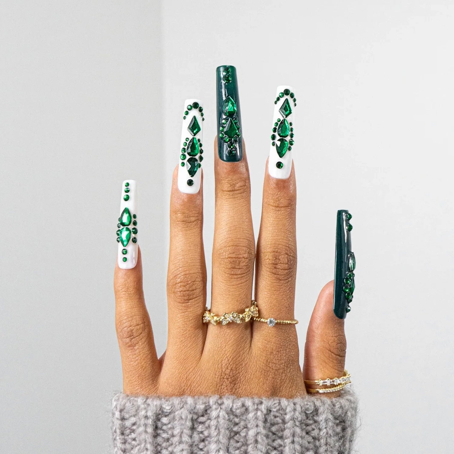 A hand with long coffin-shaped nails decorated with green and white nail art, wearing gold rings, against a light gray background.