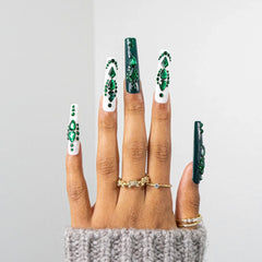 A hand with long coffin-shaped nails decorated with green and white nail art, wearing gold rings, against a light gray background.