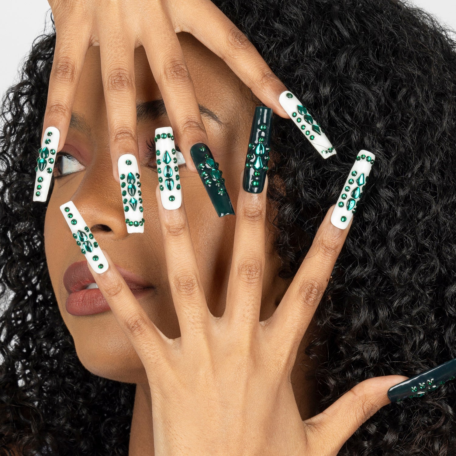 A close-up of a person's hand with long coffin-shaped nails decorated in white and dark green patterns, posed in front of their face with curly black hair.