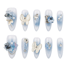 Ten false press-on nails arranged in two rows of five on a plain white background. The nails have a pale icy blue to white ombré finish and are decorated with various embellishments including white butterflies, blue crystals, pearls, and metallic charms.