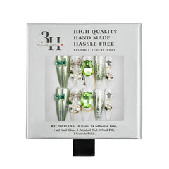 A boxed set of decorative press-on nails in a white retail package. The nails are long and embellished with green gem-like accents, silver rhinestones, bows, and metallic details.