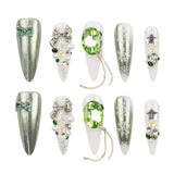 Eight artificial nail tips arranged in two rows on a white background, decorated with silver glitter, white snowflakes, bows, pearls, green gems, and star-shaped rhinestones.