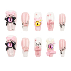 A set of ten decorative press-on nails arranged in two rows on a plain white background. The nails feature pastel pink and white glossy bases with various embellishments, including pink bows, black spider-like claw accents with purple and pink eyes, bunny ears, script text, and a small white flower.