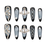 A set of 10 coffin-shaped press-on nails arranged in two rows of five on a plain white background. The nails feature a black and gray smoky gradient base with various silver and white decorative designs, including a snake, a ring-like planet, geometric lattice patterns, and faux pearl details.