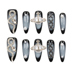 A set of 10 coffin-shaped press-on nails arranged in two rows of five on a plain white background. The nails feature a black and gray smoky gradient base with various silver and white decorative designs, including a snake, a ring-like planet, geometric lattice patterns, and faux pearl details.