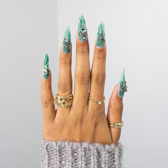 A close-up of a hand with long, sculpted stiletto nails painted in a glossy green and white marbled pattern with silver rhinestone accents. The hand is styled with multiple gold rings and a gray knit sweater cuff, set against a clean, light gray background.