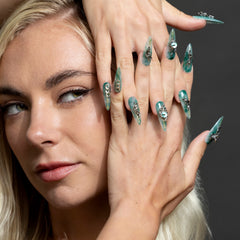A close-up of a woman's face and hands showcasing long, stiletto-shaped false nails with a glossy mint green and silver glitter design and rhinestone embellishments.