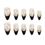 Ten false press-on nails arranged in two rows on a plain white background. Each nail has a glossy black pointed toe and a pale blush-beige upper section decorated with small gold foil flakes and rhinestone clusters.