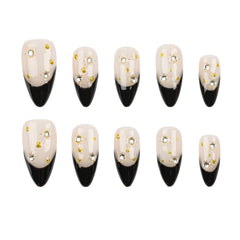 Ten false press-on nails arranged in two rows on a plain white background. Each nail has a glossy black pointed toe and a pale blush-beige upper section decorated with small gold foil flakes and rhinestone clusters.