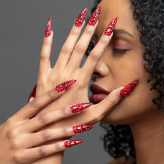 A close-up of a model's hands showcasing long, stiletto-shaped nails decorated with a red glitter and rhinestone pattern, set against a neutral gray background.