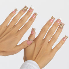 A pair of hands displaying a French manicure with pink press-on nails, gold glitter accents, and rhinestone embellishments against a light gray background.