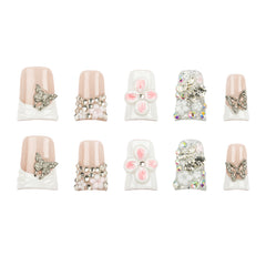 Ten decorative press-on nails arranged in two rows on a plain white background. The set includes nude pink and white nails embellished with rhinestone butterflies, pearl bead wraps, pink flower centers, and clear rhinestone clusters.