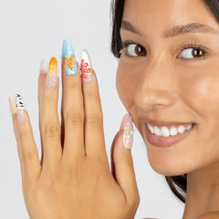 A close-up of a woman's face and hand displaying colorful, decorated press-on nails against a light gray background.