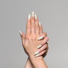 A pair of hands with long almond-shaped nails painted in white with small black polka dots, posed against a solid gray background.